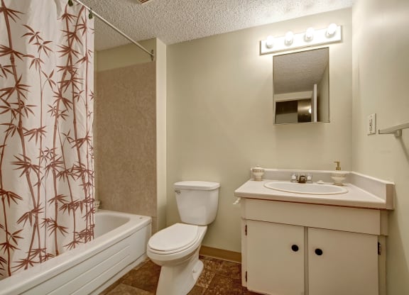 Compact bathroom here at Lakewood Heights Apartment Homes with beige walls, featuring a bamboo-patterned shower curtain, white toilet, and sink with a mirror above. Soft, warm lighting.