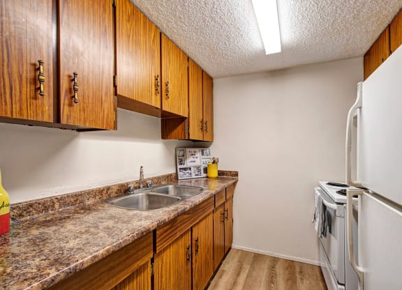 Narrow kitchen here at Lakewood Heights Apartment Homes with brown wood cabinets, marble countertops, a double sink, and white appliances. Bright, clean, and simple atmosphere.