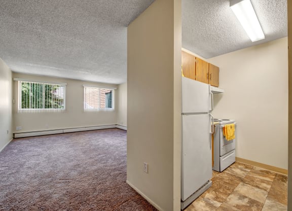 Spacious empty room here at Lakewood Heights Apartment Homes with beige carpet and two large windows with vertical blinds. Adjacent kitchen with white appliances and yellow towel. Bright and minimal.