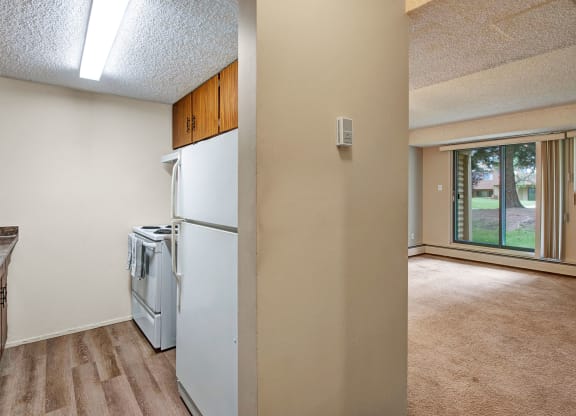 Compact kitchen here at Lakewood Heights Apartment Homes with wood cabinets and a white stove, adjacent to a bright living room with beige carpet and large window showing a grassy view. Cozy atmosphere.