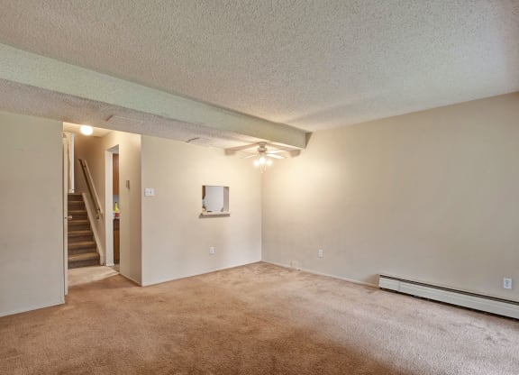 Empty living room here at Lakewood Heights Apartment Homes with beige walls and carpet, ceiling fan, and a small window opening to a kitchen area. Stairs are visible in the background.