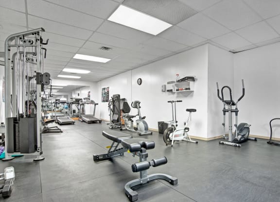 Gym interior here at Marquis Towers Apartment Homes with various exercise equipment, including treadmills, stationary bikes, and weight benches, in a clean, well-lit space with a white ceiling.
