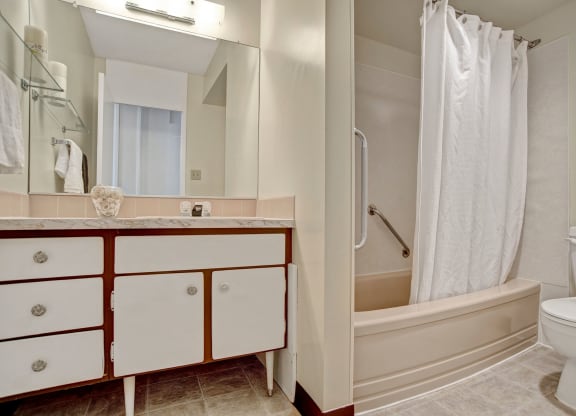 A clean bathroom here at Marquis Towers Apartment Homes with a cream bathtub and white shower curtain. A white toilet is nearby. The vanity features marble countertops and large mirrors, creating a fresh ambiance.