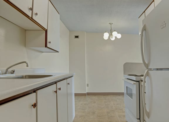Small, minimalist kitchen here at Marquis Towers Apartment Homes with white cabinets, a stainless steel sink, white appliances including a fridge and stove, and a ceiling light fixture.