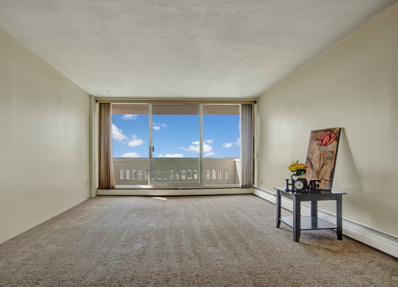 Empty room here at Marquis Towers Apartment Homes with beige carpet and walls, featuring a large glass door opening to a balcony. A small table with a floral picture and "Home" decor sits to the right.