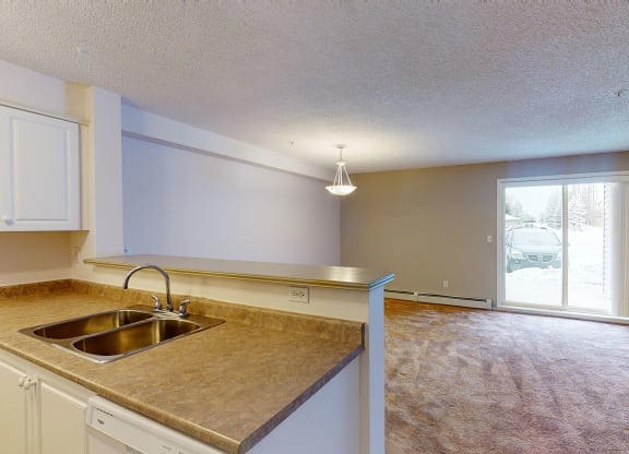 Spacious living area here at The Pointe at Applewood with beige carpet, light purple walls, and a central hanging light. A kitchen counter with a sink is in the foreground. A glass door leads outside to a snowy scene. Cozy and inviting atmosphere.