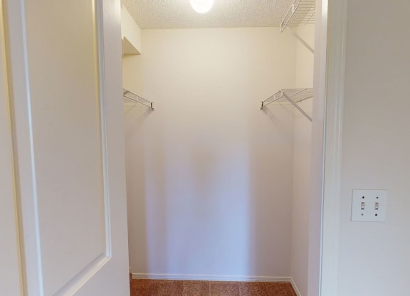 Empty closet here at The Pointe at Applewood with beige walls and a beige carpeted floor, illuminated by a ceiling light. Metal racks are mounted on the left and right sides.