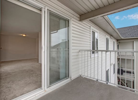 View of a small balcony here at The Pointe at Applewood with white railing attached to a white building, leading to an empty room with beige carpet and a ceiling light.