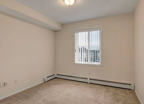 Empty room here at The Pointe at Applewood with beige walls and carpet, brightened by a single window with vertical blinds. The room has a ceiling light and baseboard heaters.