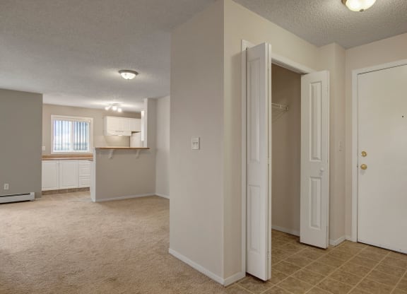 Empty apartment interior here at The Pointe at Applewood with beige carpet and tile flooring. Open closet and white front door on the right, leading to a bright kitchen with white cabinets.