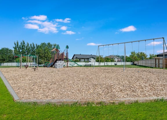 Playground here at Sol Terrace Apartment Homes with swings and climbing set, surrounded by wood chips and grass. Houses and trees in the background under a bright blue sky.