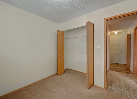 Empty room here at Sol Terrace Apartment Homes with beige carpet, open wooden closet doors revealing a shelf, and an entrance leading to a lit hallway with a closed white door. Simple and neutral atmosphere.