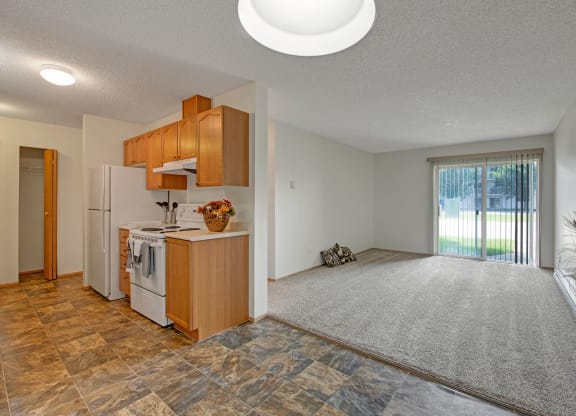 Modern open-plan apartment here at Sol Terrace Apartment Homes with brown tile kitchen, oak cabinets, and carpeted living area. Sliding glass doors lead to a bright, grassy yard.