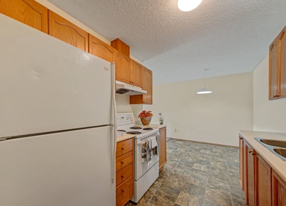 Simple kitchen here at Sol Terrace Apartment Homes with wooden cabinets and gray-tiled floor. Features a white fridge, stove, and a countertop with a basket of flowers, under warm lighting.