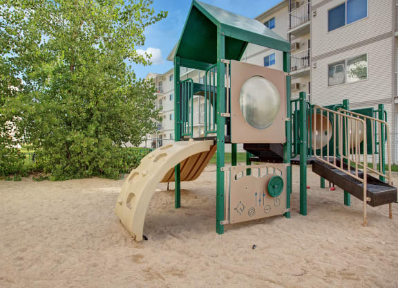 Playground scene here at Villagio Apartment Homes with a green and beige slide set on sandy ground. Lush trees in the background and residential buildings create a serene, inviting atmosphere.