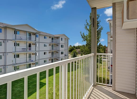View from a balcony here at Villagio Apartment Homes with white railing, overlooking a grassy area and a multi-story apartment building under a clear blue sky. Peaceful atmosphere.