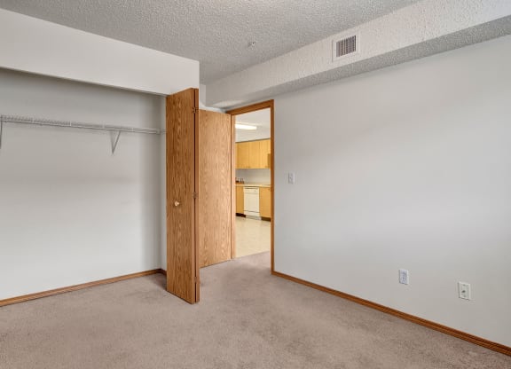 Empty room here at Villagio Apartment Homes with beige carpet, white walls, and an open wooden closet. A door leads to a kitchen with light wood cabinets, creating a neutral, open tone.