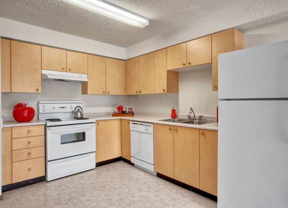 A bright kitchen here at Villagio Apartment Homes with light wood cabinets, white appliances, and a tiled floor. Red accents, like a kettle and cookware, add a touch of color and warmth.