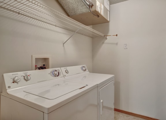 Small laundry room here at Villagio Apartment Homes with a white washer and dryer, white wire shelving above, and neutral walls. The space is clean and organized, conveying simplicity.