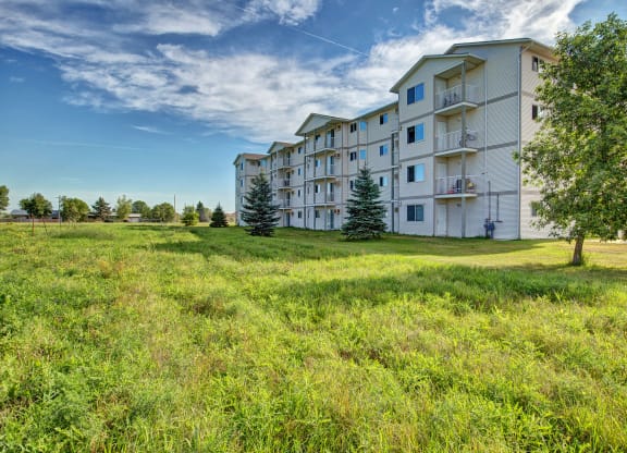 Four-story white Villagio Apartment Homes building with balconies on a sunny day, surrounded by a lush grassy lawn and trees. Blue sky with scattered clouds.