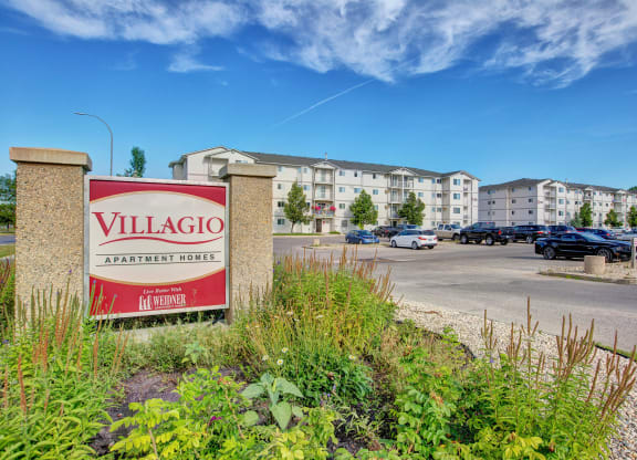 Sign reads "Villagio Apartment Homes" in front of a modern apartment complex with a parking lot and landscaped greenery under a clear blue sky.