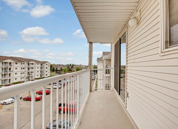 A beige apartment balcony here at Westhaven Estates Apartment Homes with white railings overlooks a parking lot and nearby buildings. The sky is blue with scattered clouds, creating a calm ambiance.