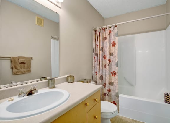 A clean bathroom here at Westhaven Estates Apartment Homes with a neutral color scheme features a white sink set in a light countertop, a large mirror, and a floral-patterned shower curtain.