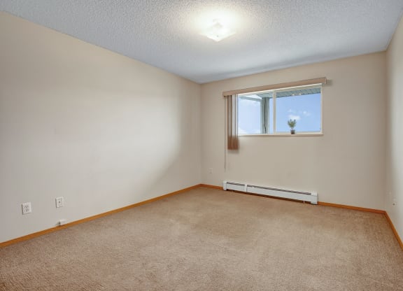 Empty room here at Westhaven Estates Apartment Homes with beige walls and carpet, a large window showing a clear blue sky, vertical blinds, and a small plant on the windowsill. Calm and neutral tone.