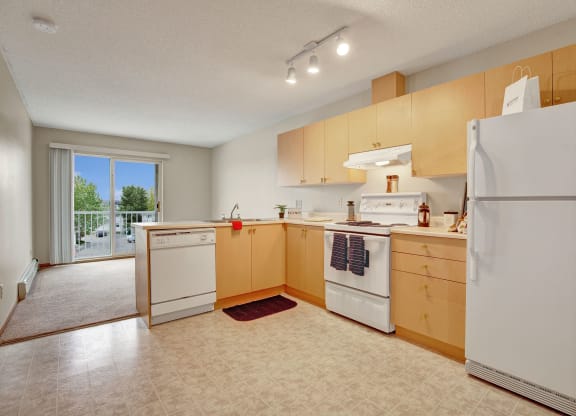 Bright kitchen here at Westhaven Estates Apartment Homes with light wood cabinets, white appliances, and under-cabinet lighting. Sliding glass doors lead to a balcony with a tree view.