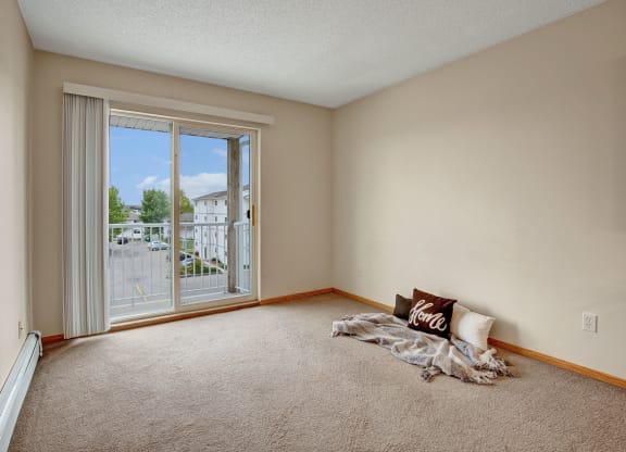 Empty room here at Westhaven Estates Apartment Homes with beige walls and carpet, featuring a sliding glass door leading to a balcony. A cozy corner has throw pillows and a blanket, conveying a welcoming feel.