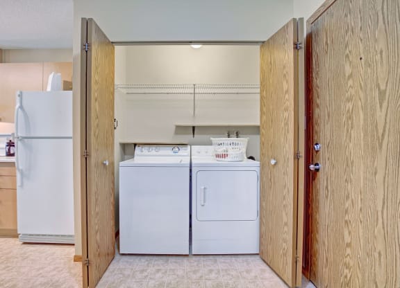 Laundry area here at Westhaven Estates Apartment Homes with wooden doors opened, revealing a white washer and dryer set. A wire shelf above holds a laundry basket. Adjacent kitchen features a fridge. Clean and organized space.