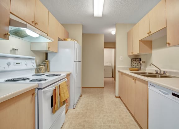 A bright, narrow kitchen here at Westhaven Estates Apartment Homes with light wood cabinets, a white stove and fridge on the left, and a dishwasher and double sink on the right. A cozy, inviting atmosphere.