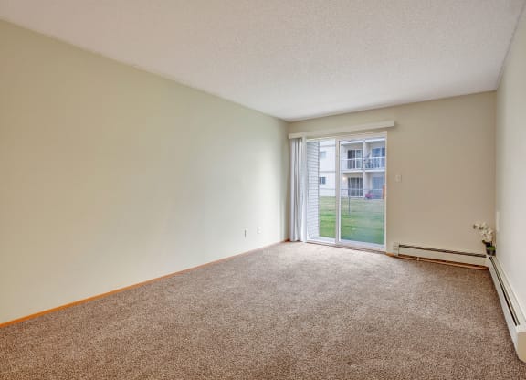Empty living room here at Westhaven Estates Apartment Homes with beige carpet, light walls, and large sliding glass doors opening to a grassy courtyard. A small plant is by the wall, creating a calm atmosphere.