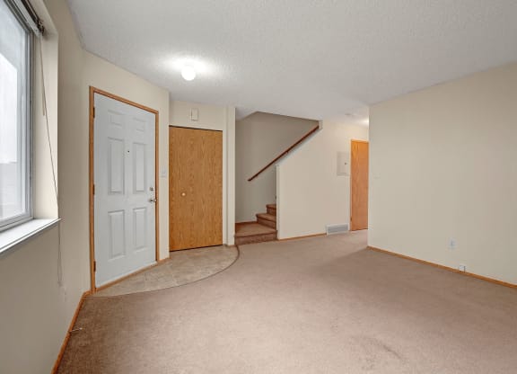 Empty living room  here at Westhaven Estates Apartment Homes with beige carpet, white walls, and a window on the left. A door and closet are near carpeted stairs leading upward, creating a calm and neutral setting.