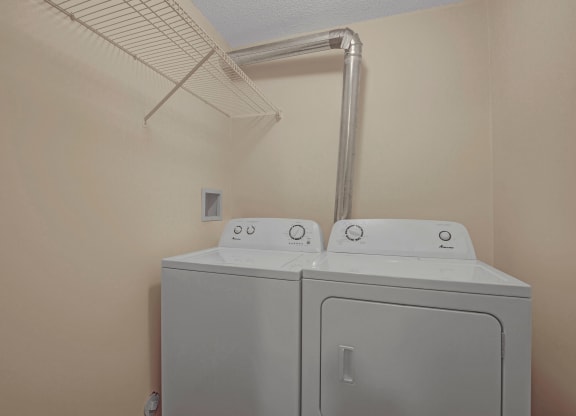 Lonely laundry room here at Westhaven Estates Apartment Homes with beige walls, white washer and dryer side by side, metal vent pipe above, and a wire shelf on the left wall.