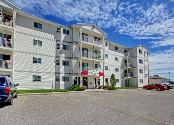 Four-story white Westhaven Estates Apartment Homes building under a vibrant blue sky with scattered clouds. The entrance has a maroon awning and there are parked cars in front.
