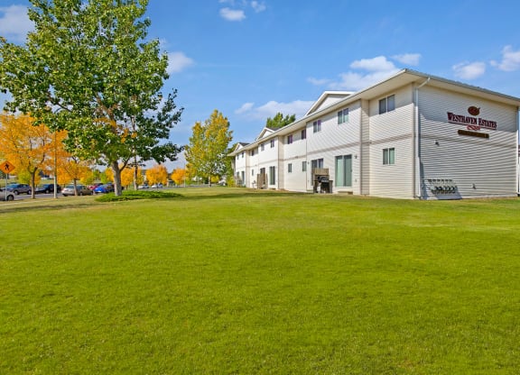 Apartment building labeled "Westhaven Estates" with white exterior, surrounded by green lawn, trees with autumn leaves, and bright blue sky.