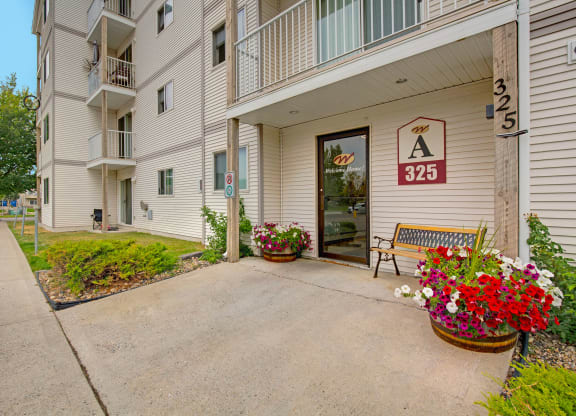Entrance of a beige apartment building here at Westhaven Estates Apartment Homes with a "325" sign. Bright flower pots and a bench create a welcoming atmosphere. Pathway and trees are visible.