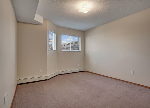Empty room here at Windsor Terrace Apartment Homes with beige walls and carpet, and two windows showing blue sky. A ceiling light illuminates the space with a calming ambience.