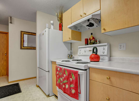 Cozy kitchen here at Windsor Terrace Apartment Homes featuring a white stove with red teapot and a red towel adorned with vegetable motifs. Light wooden cabinets add warmth.
