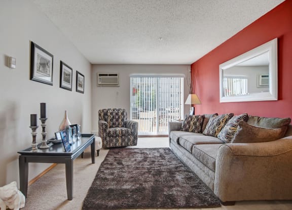 Cozy living room here at Windsor Terrace Apartment Homes with a gray couch and patterned armchair on beige carpet. A red accent wall features a large mirror. Soft light filters through vertical blinds.