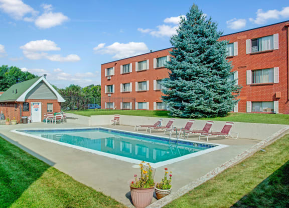 Bristol Square Apartment Homes complex with red brick buildings and a serene pool area. The pool is surrounded by lounge chairs and a large evergreen tree, conveying a relaxing atmosphere.