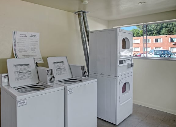 A laundromat here at Bristol Square Apartment Homes with two top-loading washers and a stacked dryer. Instruction sheets hang on the wall, and a window shows parked cars and a building.