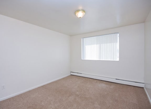 Empty room here at Bristol Square Apartment Homes with white walls, beige carpet, and a ceiling light fixture. A window with vertical blinds allows natural light, creating a neutral, calm tone.