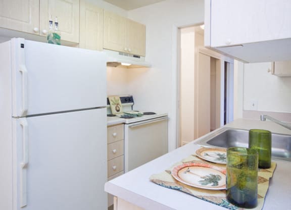 A bright kitchen here at Bristol Square Apartment Homes with light wood cabinets, a white fridge, and a stove. Counter features two green glasses, plates with leaf designs, conveying a fresh, modern vibe.