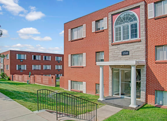 Red brick Bristol Square Apartment Homes buildings under a blue sky, with large windows and white trim. A grassy area and sidewalk lead to a columned entrance marked "1510".