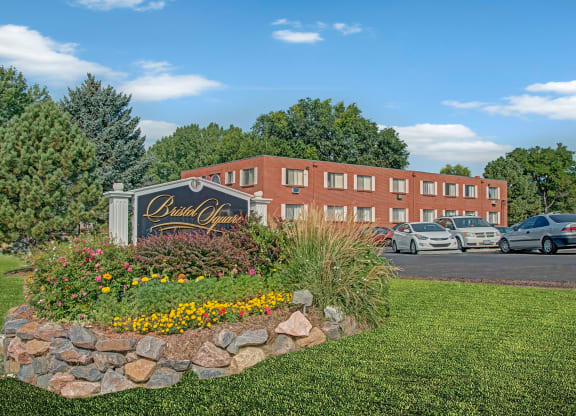 Manicured garden with colorful flowers and a "Bristol Square" sign in front. A brick residential building with parked cars is visible behind, under clear blue sky.