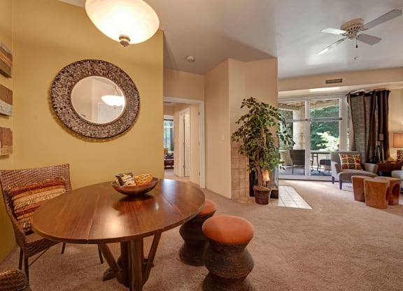 Stylish apartment interior here at Cheyenne Creek Apartment Homes with a circular dining table, wicker chairs, wall mirror, and potted plant. Bright, cozy vibe with large windows and light fixture.