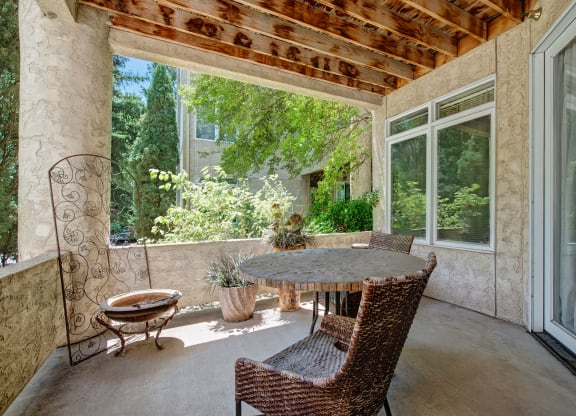 A serene patio here at Cheyenne Creek Apartment Homes with a rustic round table and two wicker chairs. Sunlight filters through overhead beams, and lush greenery surrounds the space, creating a tranquil ambiance.
