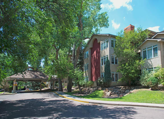 A residential building here at Cheyenne Creek Apartment Homes with large windows is surrounded by lush trees and greenery under a clear blue sky. The scene is serene and inviting.